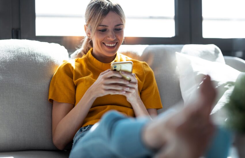 Pretty young smiling woman using mobile phone sitting on a couch at home